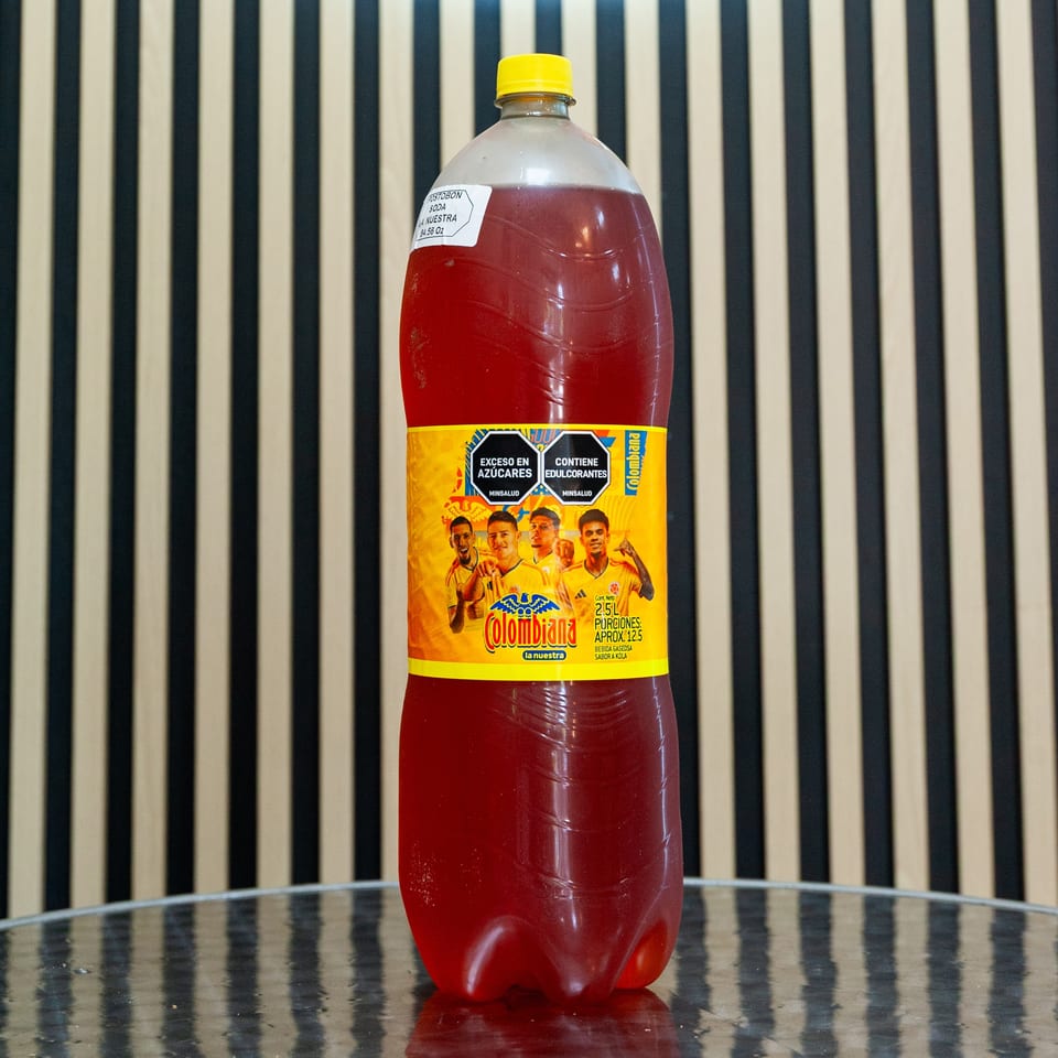 Large bottle of red Colombiana soda with a yellow cap on a reflective table against a striped wall.