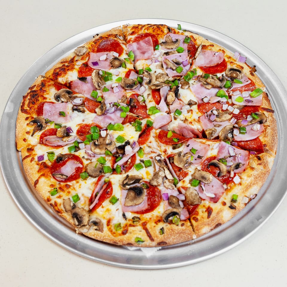 Round pizza topped with pepperoni, ham, mushrooms, green peppers, onions, and melted cheese on a metal tray.