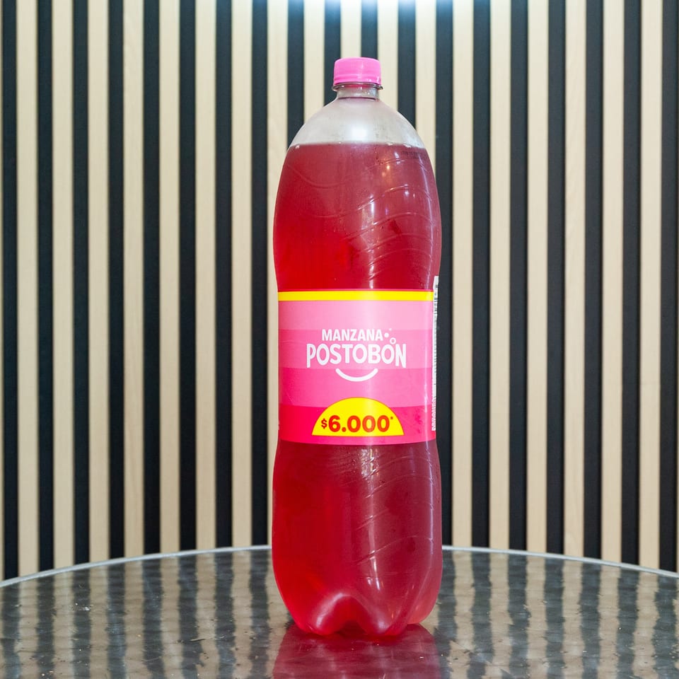 Large bottle of red Postobón Manzana apple soda with a pink cap and $6,000 price label on a reflective table.
