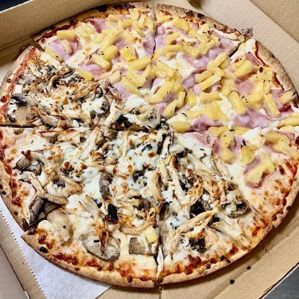 Large thin-crust pizza in a cardboard box, half topped with ham and pineapple and half with mushrooms, onions, and melted cheese.