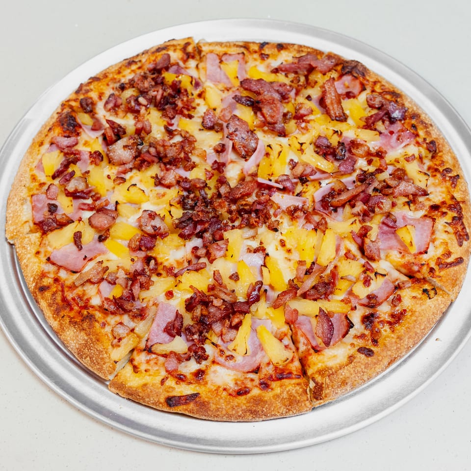 Whole Hawaiian pizza topped with ham, pineapple, bacon, and melted cheese on a metal serving tray.