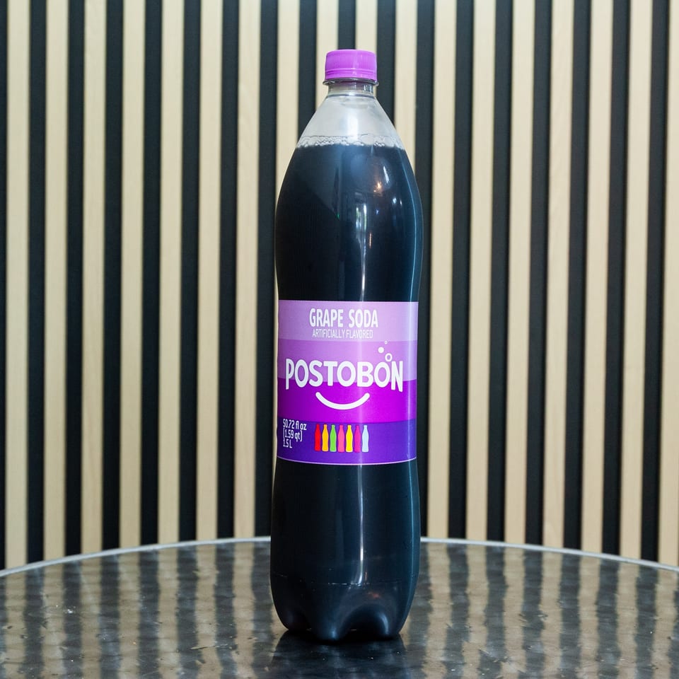 Bottle of Postobón artificially flavored grape soda on a reflective table against a striped black-and-cream background.