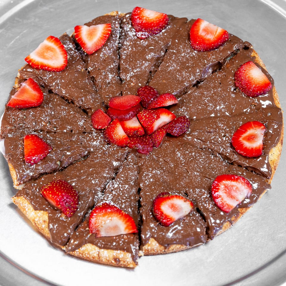 Sliced chocolate dessert pizza topped with powdered sugar and fresh strawberries on a metal serving tray.